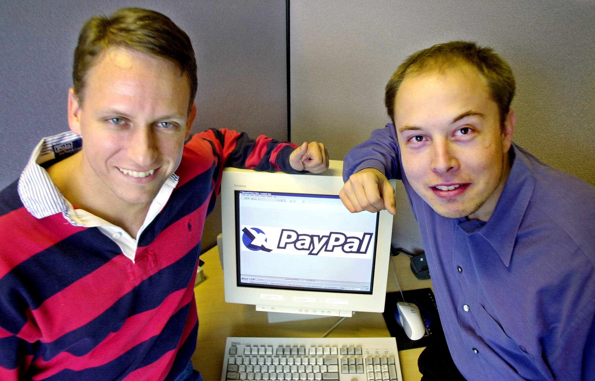 Peter Thiel and Elon Musk beside a desktop monitor displaying the early PayPal logo.